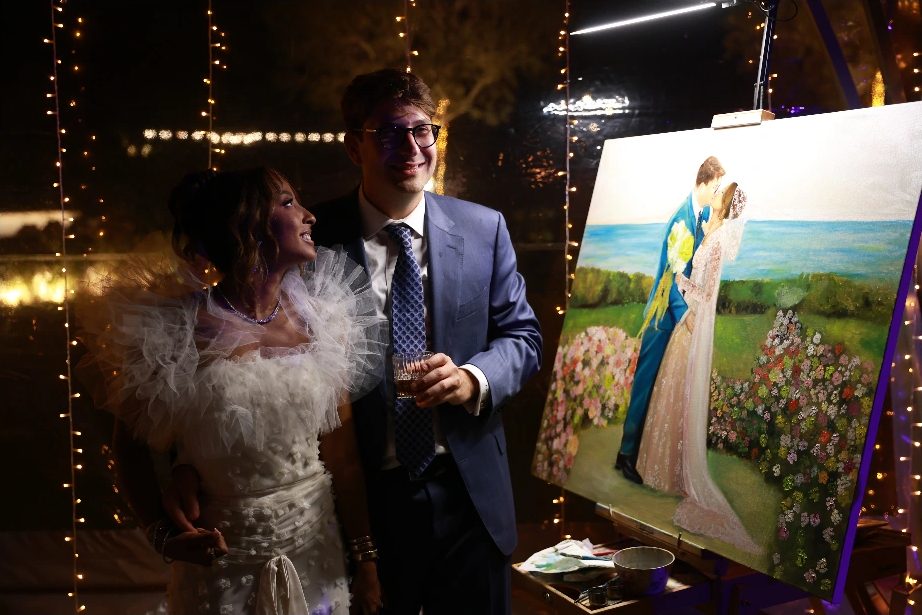 Giannis and Alexandria smiling beside their large live wedding painting at night at Island, Athens Riviera