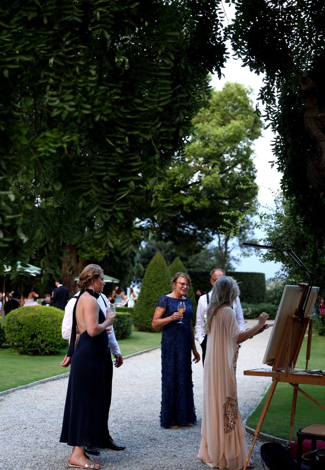 Guests chatting with live wedding painter Angela Yfantidou at her easel during the aperitivo at Villa Pizzo